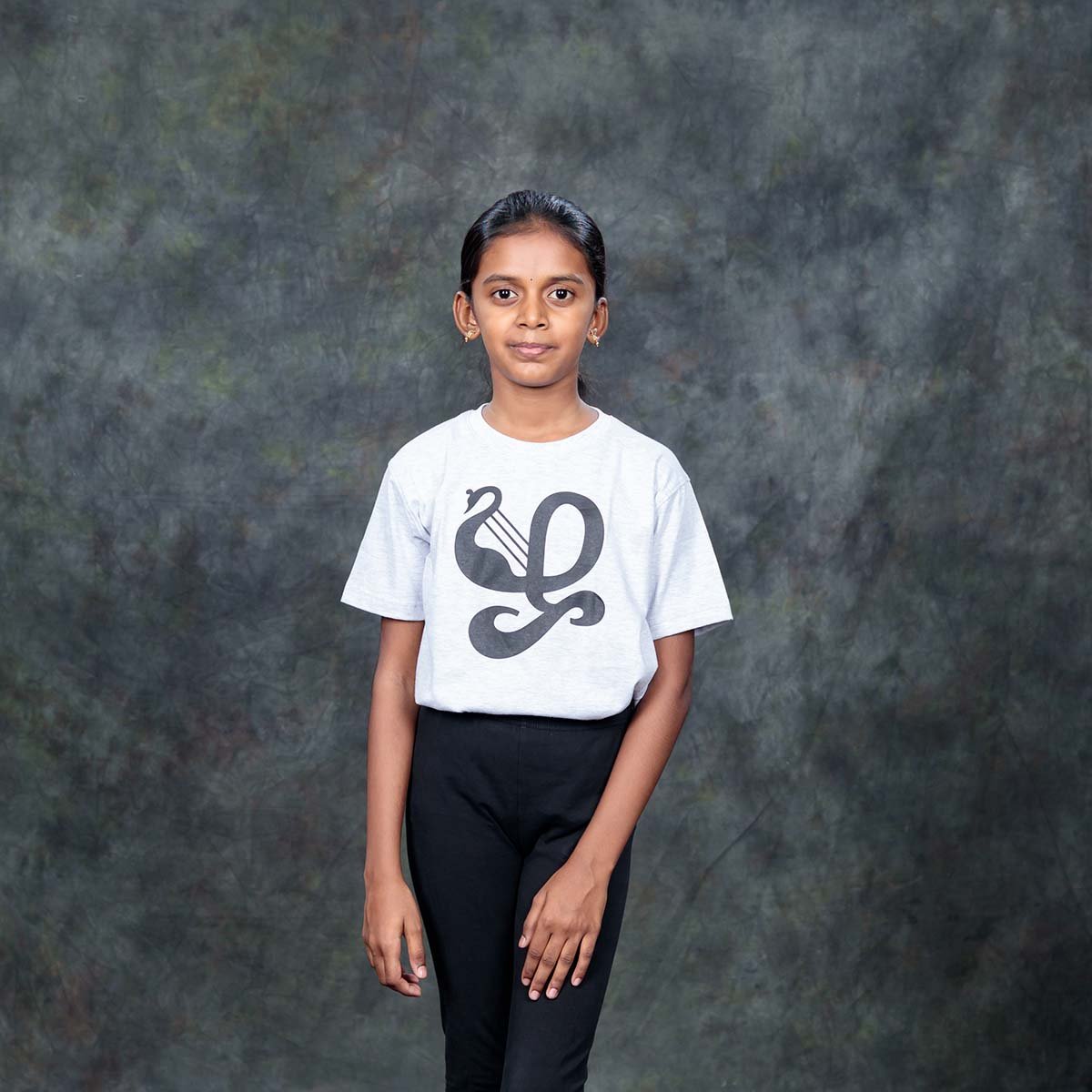 A young girl standing full length in an indoor studio, modeling a kids ecru Tamil identity t-shirt featuring the black Zha typography design.