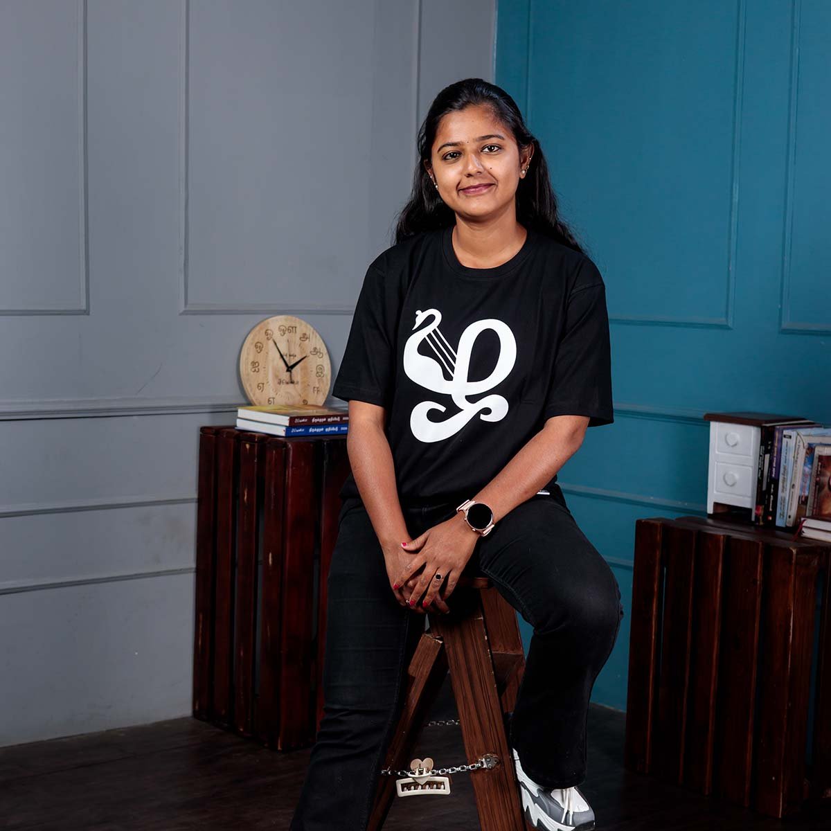 Adult female model smiling while seated on a wooden stool indoors, wearing a black Venbaa Zha typography t-shirt with a bold white graphic print.