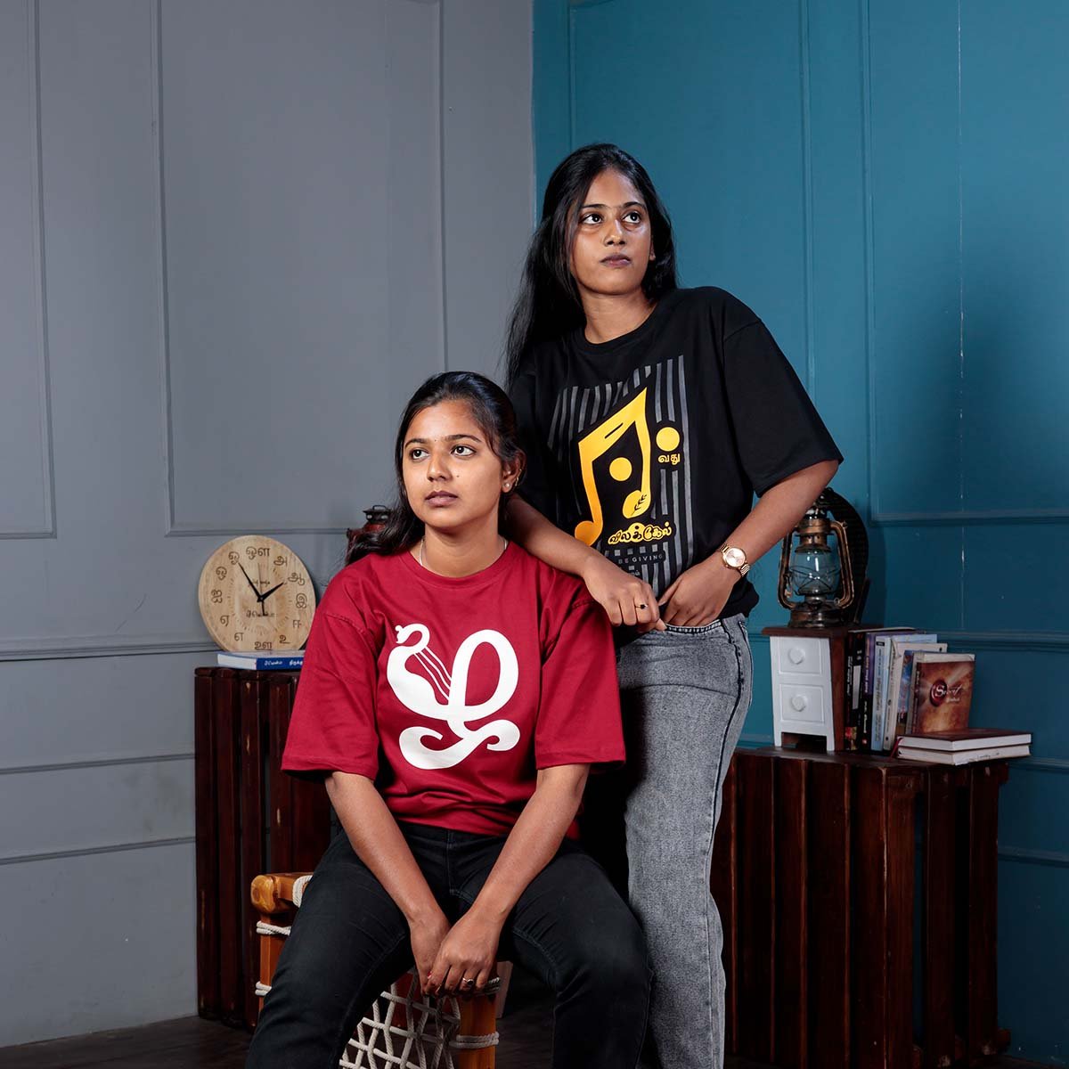 Two female models posing indoors; one seated model wears the maroon Venbaa Zha typography t-shirt while the standing model wears the black Eevathu Vilakel tee.