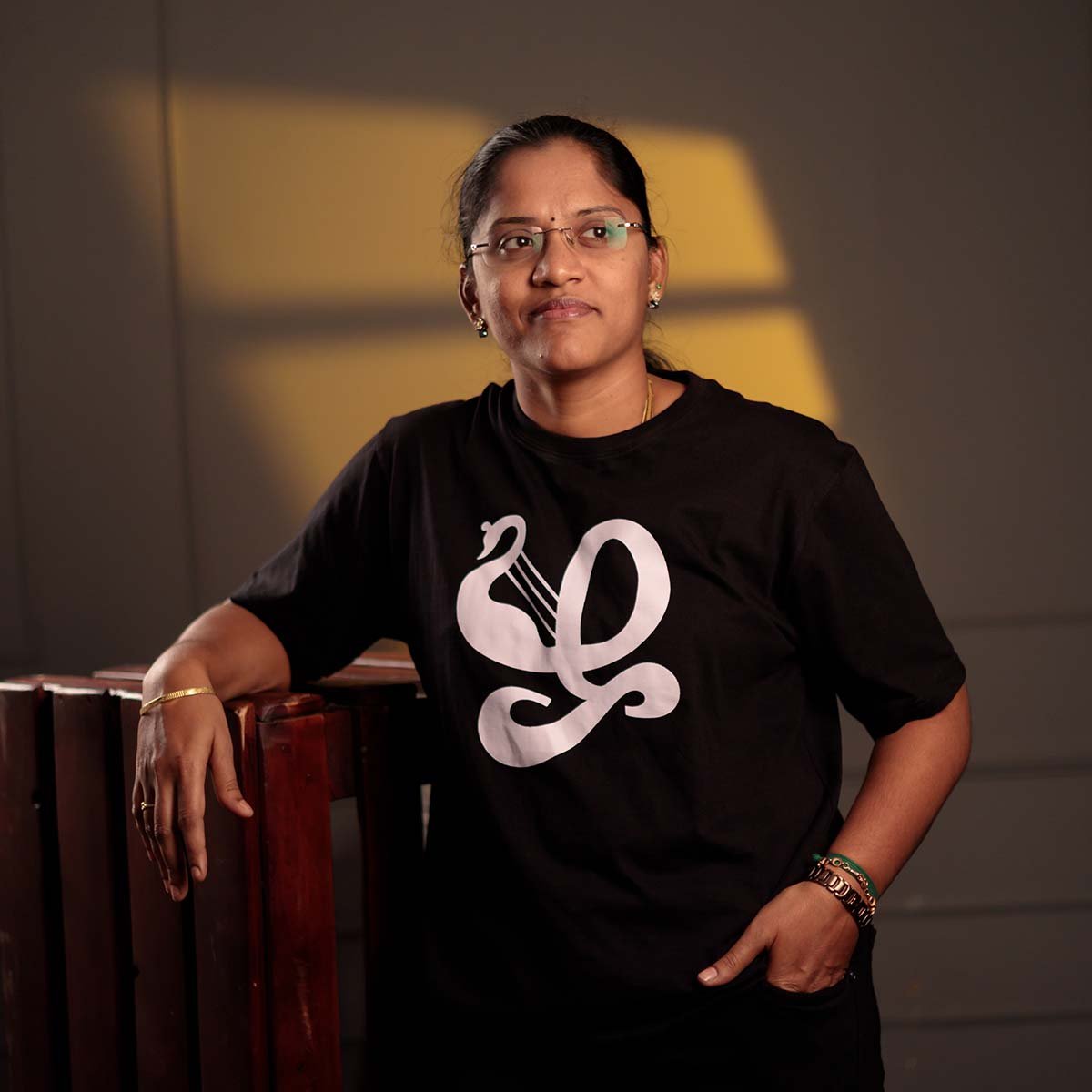 Adult female model wearing a black Venbaa Zha typography t-shirt while leaning on a wooden structure in an indoor studio with dramatic warm window lighting