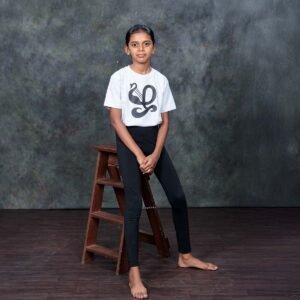 Older child model sitting gracefully against a wooden ladder stool, showcasing the clean fit of the ecru kids Zha typography t-shirt against a textured grey backdrop