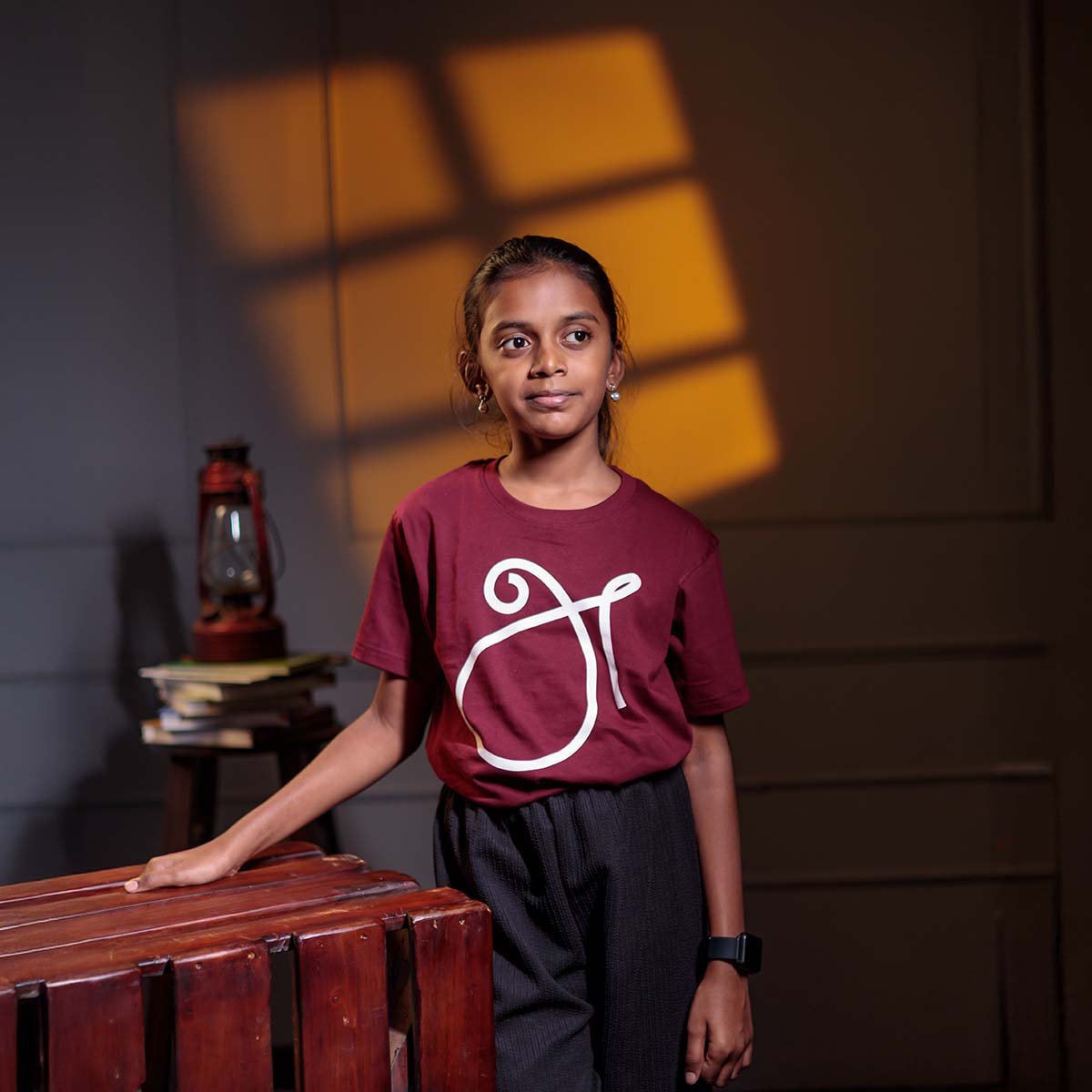 Older child model leaning on a wooden structure, showcasing the kids version of the maroon Tamil First Letter 'அ' (உயிர் எழுத்து) typography t-shirt.