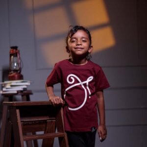 Younger child model smiling while leaning on a small wooden ladder, wearing the kids maroon 'அ' Tamil First Letter (உயிர் எழுத்து) graphic t-shirt.