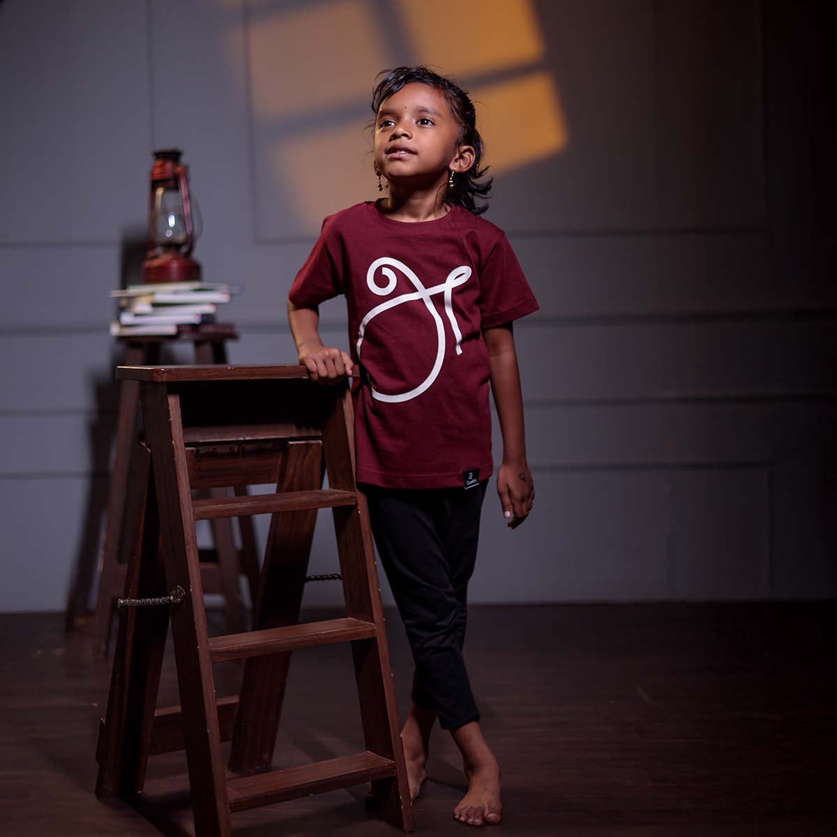 Full-length view of a young girl standing next to a wooden ladder step-stool, modeling the maroon kids 'அ' Tamil First Letter script t-shirt.