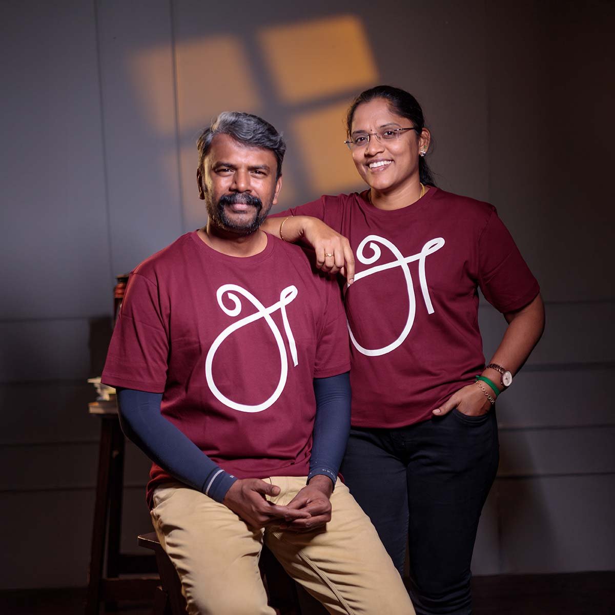 dult male and female models smiling together in an indoor studio, wearing matching maroon 'அ' Tamil First Letter (உயிர் எழுத்து) identity t-shirts.