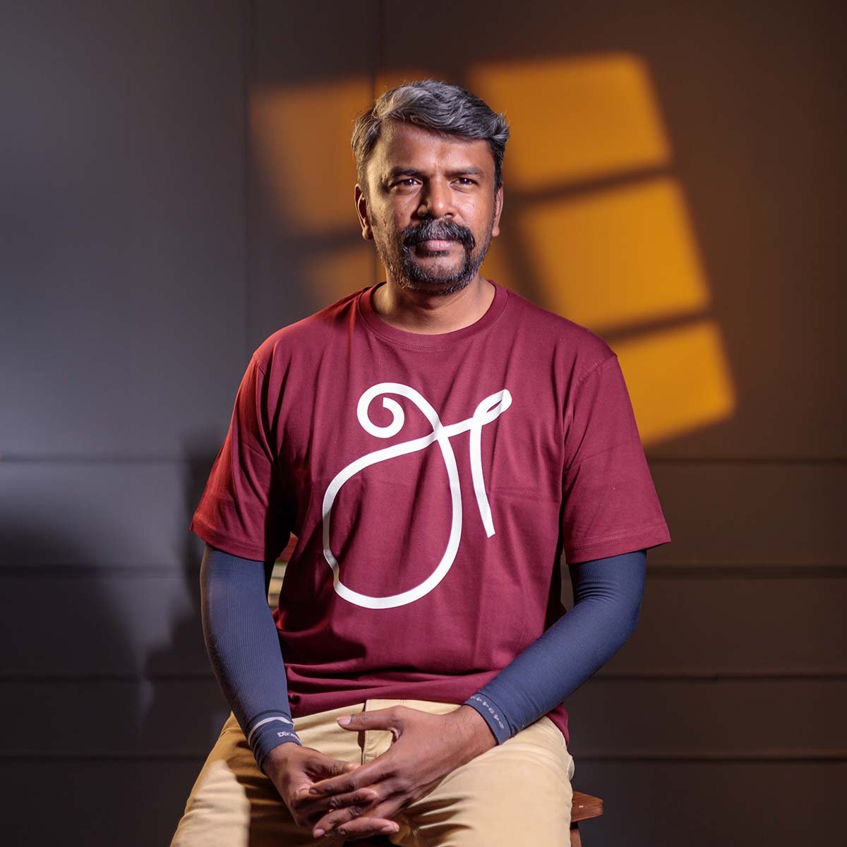 Adult male model with a mustache sitting down, wearing the maroon 'அ' First Letter (உயிர் எழுத்து) typography t-shirt under dramatic studio window lighting.