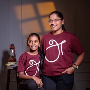 Smiling mother and daughter posing closely together indoors, wearing matching maroon 'அ' Tamil First Letter identity t-shirts.