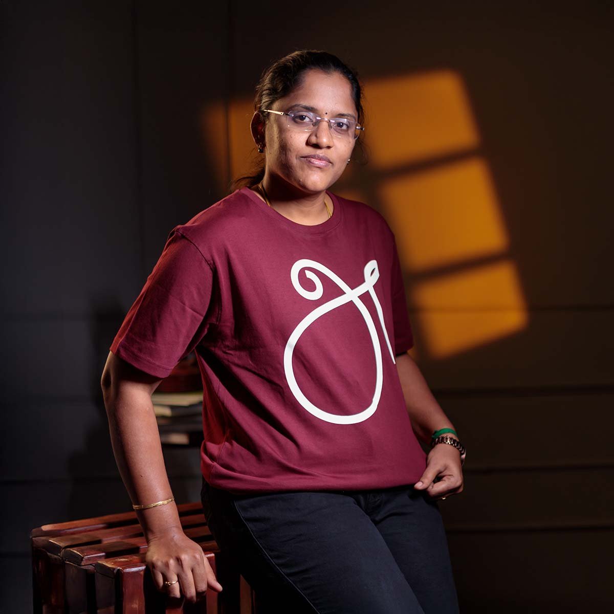 Adult female model wearing glasses looking to the side, showcasing the maroon unisex Tamil First Letter 'அ' (உயிர் எழுத்து) print t-shirt in a studio setting.