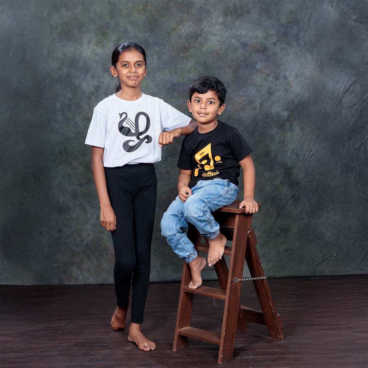 An older girl wearing the ecru Zha typography t-shirt standing next to a young boy seated on a wooden ladder stool wearing the black and golden yellow Eevathu Vilakel shirt
