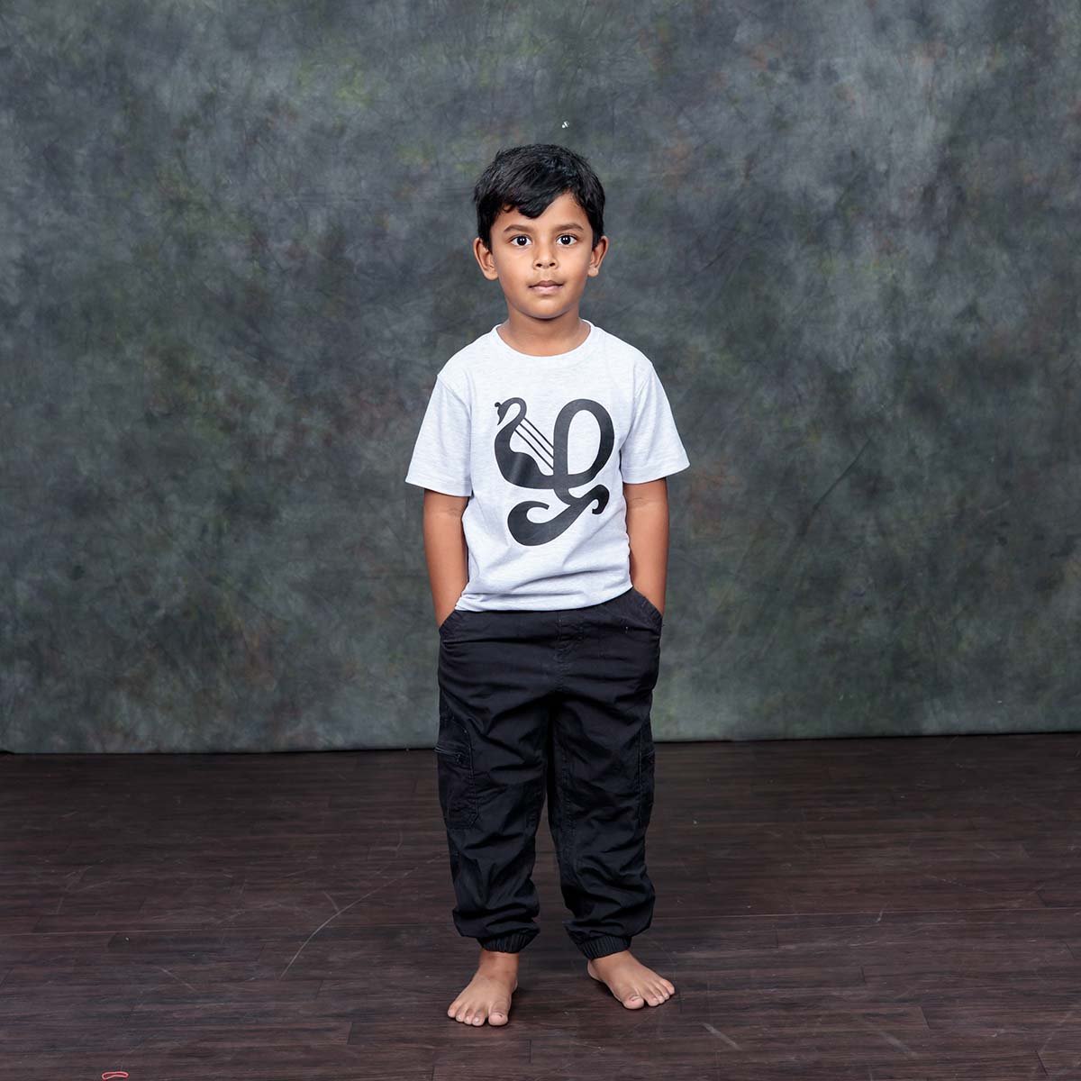Young boy standing barefoot with hands in his pockets, modeling the ecru off-white kids Zha typography t-shirt against a textured dark grey studio backdrop.