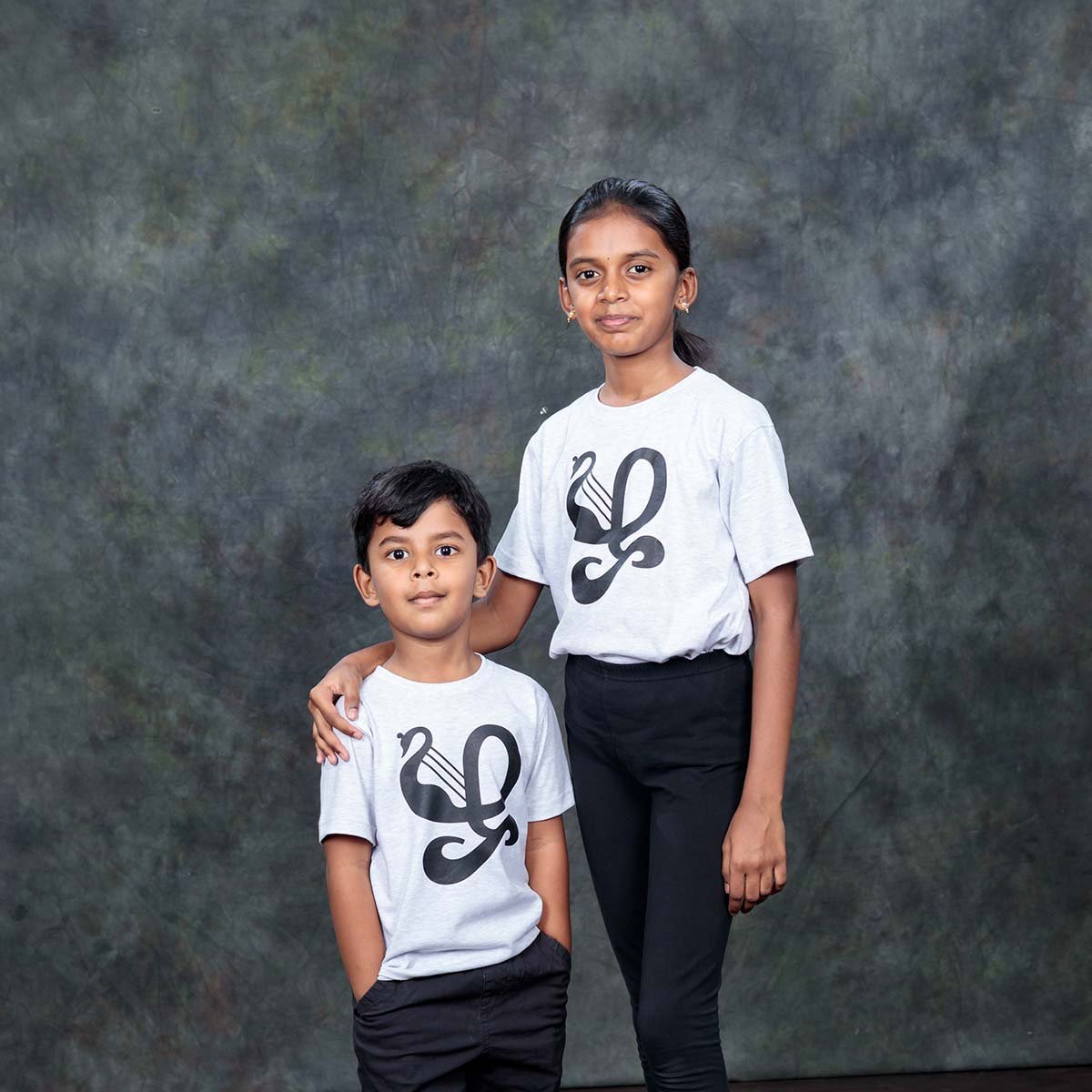 Older girl and younger boy posing tightly together in an indoor studio, wearing matching ecru kids Zha typography printed t-shirts.