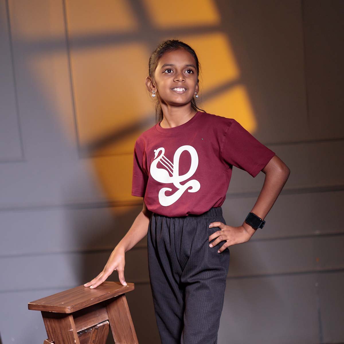 Older child model standing confidently with her hand on her hip, showcasing the tucked-in maroon kids Zha typography t-shirt against a grey wall with warm lighting shadows.