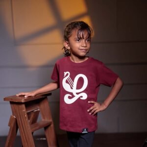 Younger child model posing with her hand on her hip next to a wooden stool, wearing the deep maroon kids Zha typography t-shirt under warm studio lighting