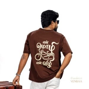 Man wearing a brown oversized Venbaa t-shirt with a cream 'என் மொழி என் வழி' motorcycle typography print on the back standing against a white studio background